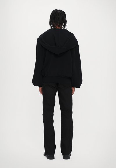 Hanorac negru oversized cu glugă ajustabilă, tiv elastic și mâneci largi, purtat împreună cu pantaloni și încălțăminte negre, văzut din spate.