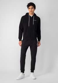 Sudadera negra con cremallera y cordones, con bolsillos frontales y logo. Combinada con joggers negros con puños. Zapatillas blancas completan el look.