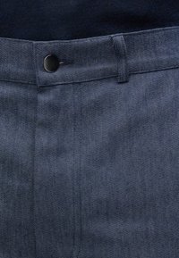 Tumši zili džinsu bikses ar pogu aizdari, plakanu priekšpusi un smalku rakstu, izdaiļo to izturīgo audumu un šūto dizainu.