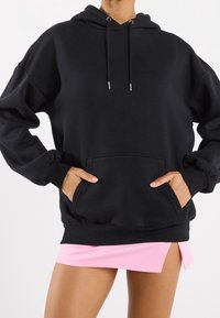 Černá oversized mikina s kapucí na stahování, přední kapsou a zpevněnými manžetami. Nositá přes světle růžovou mini sukni. Hladká textilie.