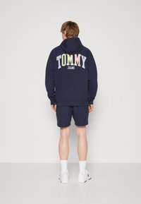 Navy hoodie with "TOMMY" in multicolor gradient text on the back, paired with matching shorts and white sneakers. Smooth fabric, casual fit.