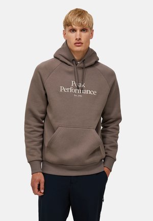 Giovane con capelli biondi che indossa una felpa marrone con cappuccio con testo "Peak Performance Est.1986" e pantaloni scuri, in piedi davanti a uno sfondo semplice.