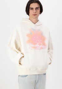 Cremfarbener Hoodie mit einem großen, pinken und blauen Grafikdesign auf der Vorderseite, einer vorderen Tasche und einer Kordelzugkapuze.