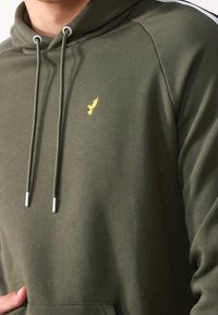 Sudadera verde olivo con cordones, puntas plateadas y un pequeño logo de un pájaro amarillo en el pecho. Tejido suave con un corte relajado.