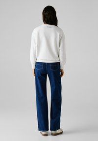 Sudadera blanca con el logo "PEPE JEANS" en la parte trasera, combinada con jeans de pierna ancha azules que cuentan con bolsillos traseros y costuras en contraste.