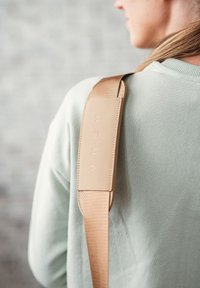 Beige verstellbarer Schulterriemen aus Stoff, mit einem glatten gepolsterten Abschnitt und geprägtem Logo. Trägt man über einem hellgrünen Kleidungsstück.