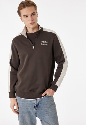 Junger Mann trägt braunen Sweatshirt mit Viertel-Reißverschluss, weißen Streifen auf den Ärmeln und dem Text "Set the Pace Embrace the Challenge" auf der Brust.