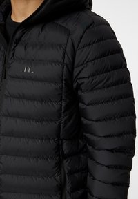 Svart puffjacka med huva, med quiltad textur, dragkedjestängning och en subtil logotyp på bröstet. Slät, lättviktsmaterial.