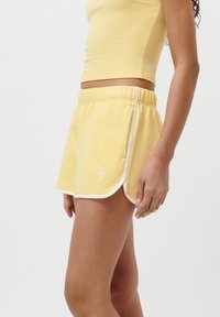 Shorts di cotone gialli con un bordo bianco, vita elastica e spacchi laterali. I pantaloni corti presentano un piccolo logo ricamato sulla parte anteriore.