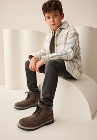 Braune Schnürstiefel aus strukturiertem Wildleder mit schwarzen Lederakzenten. Das Kind trägt ein kariertes Hemd und dunkle Jeans und sitzt auf einer geschwungenen Fläche.