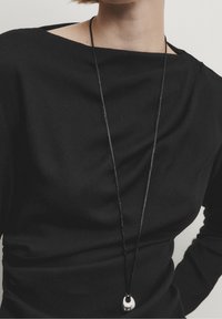 Black long-sleeve top with a boat neckline, featuring draped fabric. A black cord necklace with a silver pendant hangs visible.