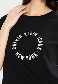 Czarna koszulka z okrągłym, białym haftowanym logo z napisem „CALVIN KLEIN JEANS NEW YORK” wyraźnie widocznym z przodu. Miękka faktura materiału.
