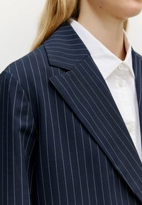 Marinblaues Blazer mit Nadelstreifen und Reverskragen, mit weißen Streifen, strukturierten Schultern und einem weißen Hemd darunter.