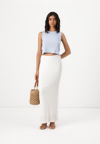 Camisola curta com riscas em azul claro, decorada com um pequeno laço, combinada com uma saia maxi branca texturizada. A modelo segura uma bolsa de palha e usa sandálias em tom de areia.