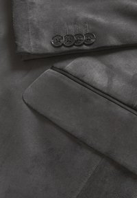 Nærbillede af ærmet på en mørkegrå suedeblazer med fire sorte knapper og en klaplomme under ærmet.