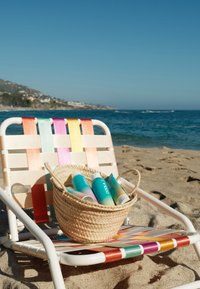Geflochtener Strandstuhl mit mehrfarbigen Bändern hält einen Korb voller Sonnencremeröhren am sandigen Ufer am Meer.