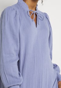 Vaaleanlila pusero pehmeästä, tekstuurillisesta kankaasta, jonka rypytys kaulus ja avainreikän muotoinen pääntie sisältävät nauhakiinnitykset ja puffihuput.