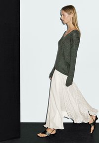 Dunkelgrüner Strickpullover, cremfarbener Maxiröcke mit einem strukturierten Karomuster, schwarze flache Sandalen, Modell läuft vor einem hellen Hintergrund.