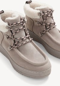 Zamszowe botki za kostkę w jasnym taupe z puszystym białym kołnierzem z futra, połączone z wzorzystymi różowo-czarnymi sznurówkami i teksturowaną gumową podeszwą.