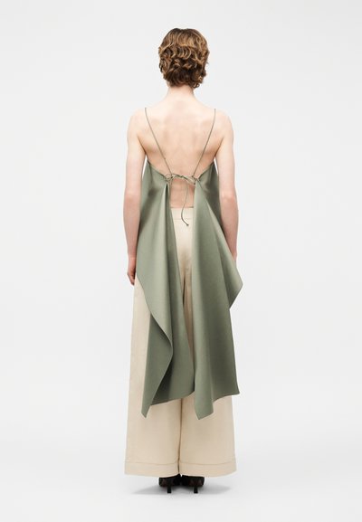 Άτομο με κοντά, σγουρά μαλλιά φοράει αμάνικο, χακί τοπ χωρίς πλάτη δεμένο στο πίσω μέρος και φαρδιά μπεζ παντελόνια, στέκεται σε ψηλοτάκουνα, με την πλάτη προς τα πίσω.