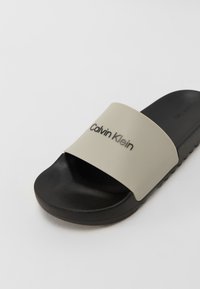 Sandale tip sliders Calvin Klein, cu o curea lată crem, pe care este imprimat numele brandului în metalizat și o talpă din cauciuc texturat negru.
