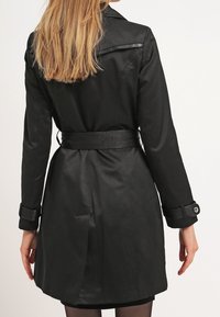 Trench-coat noir avec une taille ceinturée, des manches longues et des poignets boutonnés. Texture lisse semblable à du satin et col classique. Vue de dos.