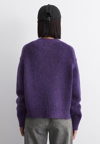 Maglione a maglia viola con una finitura testurizzata, caratterizzato da un orlo a coste e maniche ampie. Indossato con jeans grigi e un cappellino nero.
