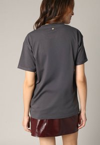 Camiseta de algodón gris con un corte relajado, cuello redondo y mangas cortas. Presenta un pequeño detalle dorado en la parte posterior, cerca del escote.