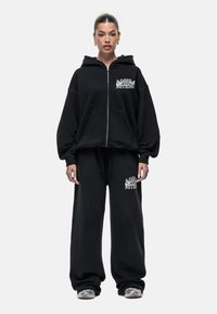Zwarte hoodie met rits in een oversized pasvorm, voorzien van een grafische "GOOD NOTHING" print. Gepaard met losse zwarte sweatpants, beide in een zachte stof.