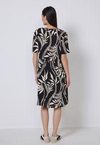 Schwarzes Kleid mit beigem Blattmuster, kurzen Ärmeln und lockerem Schnitt. Kombiniert mit lässigen Sneakers, aus glattem Stoff mit subtiler Textur.