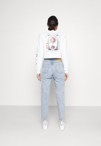 Bílý zkrácený svetr s barevnými grafickými designy na zádech a rukávech, v kombinaci se světle modrými džíny s vysokým pasem a bílými teniskami.