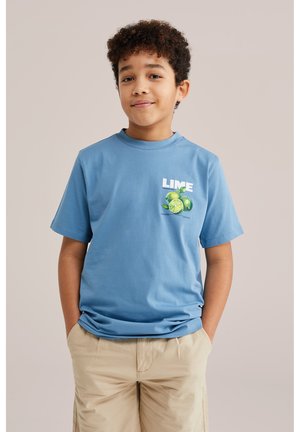 Göndör hajú fiú kék pólót visel lime mintával és bézs rövidnadrágot, zsebre tett kézzel, egyszerű háttér előtt áll.