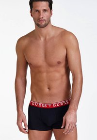 Boxeri negri, cu talie lată cu logo-ul roșu "GUESS", cu un design ajustat și textură de material neted. Modelul prezintă vederea frontală.