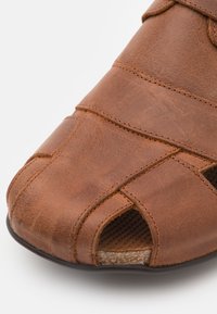 Sandal in pelle marrone con design open-toe, cinturini incrociati e finitura testurizzata. Presenta una soletta imbottita e una suola nera robusta.