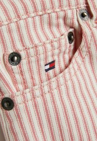 Tessuto denim con strisce verticali rosse e avorio, caratterizzato da una piccola tasca con bottoni neri e un'etichetta con logo rosso, bianco e blu.