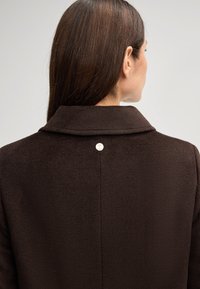 Cappotto marrone con colletto pieghevole, tessuto texturizzato e dettaglio con bottone silver sulla parte posteriore. Design semplice ed elegante, senza chiusure visibili.