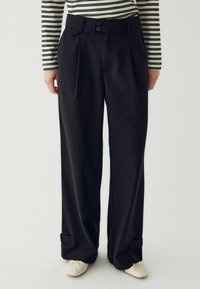 Melni platleņķa bikses ar diviem pogām, krokām un aprocēm. Gluda auduma tekstūra, kombinēta ar svītrainu garām piedurknēm topu.