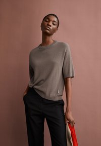 Grauer gerippter Kurzarmpullover mit Rundhalsausschnitt, kombiniert mit schwarzen Hosen. Das Modell hält einen gefalteten roten und beigen Stoff.
