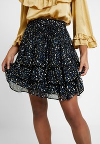 Gonna nera a strati con orlo arricciato, caratterizzata da un motivo colorato di macchie blu, oro e bianche, realizzata in materiale leggero e trasparente.