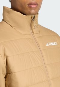 Et nærbillede af en person, der bærer en tan farvet puffy jakke med høj krave, som har Terrex-logoet på brystområdet.