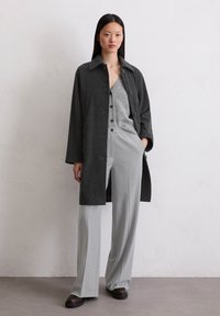 Vrouw staat met een donkergrauwe jas, lichtgrijs knopenvest, brede lichtgrijze broek en donkerenbruine schoenen tegen een witte muur.
