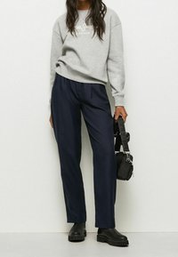 Felpa grigia con logo, pantaloni navy e stivali neri con suola spessa. Porta una piccola borsa nera con una tracolla decorata.