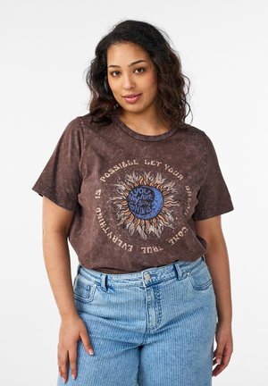 Ung kvinde iført en brun t-shirt med sol- og månegrafik samt teksten "You are my sun shine", kombineret med lyseblå jeans med højtaljede.