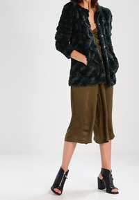 Grøn jakke i imiteret pels med tekstureret finish, knapper foran og to lommer, båret over en olivengrøn satinheldragt og sorte ankelstøvler med hæl.