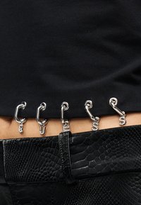 Μαύρο υφασμάτινο τοπ με διακοσμητικά ασημένια κλειδιά με λεπτομέρειες από κρυσταλλάκια. Φορεμένο πάνω από μια υφασμάτινη μαύρη ζώνη από ψεύτικο δέρμα με υφή.