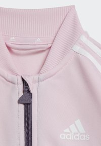 Giacca sportiva rosa con colletto a costine, chiusura zip e accenti bianchi a tre strisce. Presenta un logo adidas bianco sul davanti.