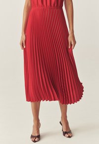 Červená plisovaná midi sukňa vyrobená z hladkej tkaniny, s priliehajúcim pásom a splývavým dizajnom, skombinovaná s hnedými sandálmi na otvorenú špičku.