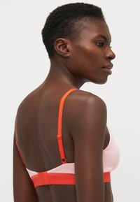 Soutien-gorge en rose et orange, doté de bretelles réglables et d'une fermeture à crochets. Le tissu a une texture lisse avec un design minimaliste.