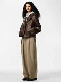 Pruun nahktagi, millel on suured kraed, allhall kapuutsiga pusa ja lõdvad beežid püksid. Modell seisab ühevärvilise tausta ees.