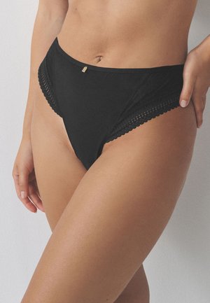 Femme portant des sous-vêtements style bikini noirs avec bordure en dentelle et petit charme doré à la taille, debout devant un fond uni.